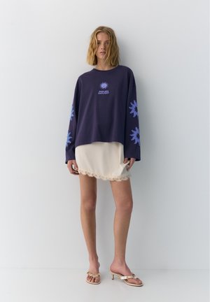 Femme debout portant un sweat-shirt oversize bleu marine avec des motifs floraux bleus et le texte "Endless Daylight", une robe nuisette crème bordée de dentelle et des sandales à talons beiges.