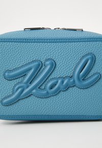 Geantă cosmetică din piele albastră, cu text embosat "Karl", suprafață texturată, accente netede și închidere cu fermoar argintie. Formă compactă, rectangulară.