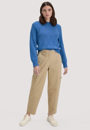 Vrouw staand, gekleed in een blauw gebreide trui, beige cargobroek en witte sneakers, tegen een lichte neutrale achtergrond.