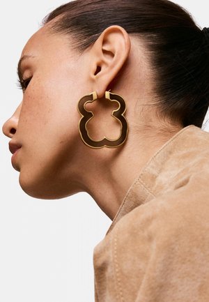 Frau mit dunkel zurückgebundenem Haar, die große goldene und dunkelbraune, abstrakt geformte Creolen und eine beige Wildlederjacke trägt.