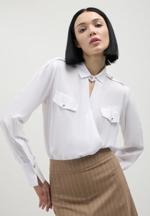 Blusa bianca a maniche lunghe con dettagli in bottoni, due tasche sul petto e scollatura a V, abbinata a una gonna beige a righe sottili. Tessuto dalla consistenza liscia.