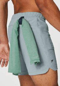 Hellgrüne Shorts mit elastischem Bund und Seitentaschen, verziert mit einem marineblauen Akzent. Ein hochgerolltes grünes Hemd ist hinten zusammengebunden.