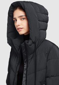 Sort puffjakke med en stor, struktureret hætte. Har lynlåslukning og diagonale syninger. Glat, tekstureret stof.