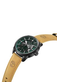 Montre en acier inoxydable noir avec un cadran transparent, des accents verts et un bracelet en cuir beige avec un logo embossé. Mécanisme squelette visible.