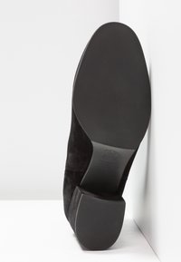 Chaussure habillée pour homme en daim noir vue par la semelle, avec une semelle extérieure en cuir lisse et un talon carré en caoutchouc sur fond blanc.