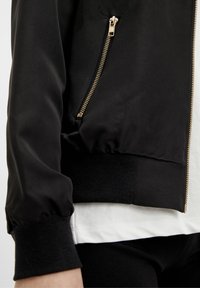 Chaqueta negra con cremallera dorada, llevada sobre una camisa blanca, mostrando la parte inferior de la manga y la cintura.