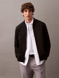 Black bomber jacket with a zip closure, round neck, and ribbed cuffs, layered over a white button-up shirt and gray trousers.