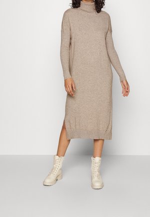 Kvinde iført en beige langærmet rullekrave strikkekjole med sideslidser, parret med off-white snørestøvler ved anklerne.