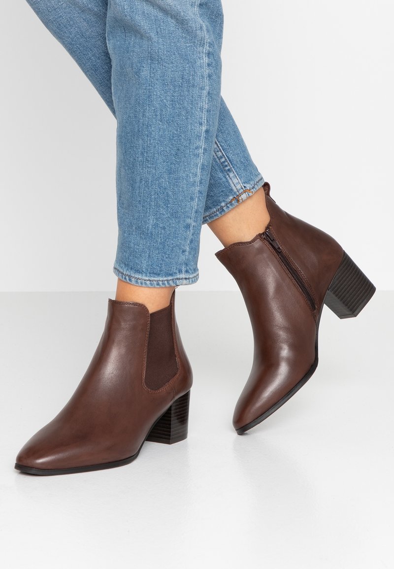 Bottes cheville en cuir marron avec bouts pointus, panneaux élastiques latéraux et talon carré. Texture lisse et détail de fermeture éclair visible.