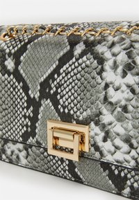 Černá a bílá kabelka s hadím vzorem, se zlatým kovovým řetízkem a čtvercovým zapínáním. Texturovaný povrch, obdélníkový tvar.