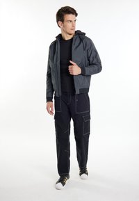 Jachetă bomber gri cu glugă, asortată cu pantaloni cargo negri cu cusături albe. Pantofi negri cu dungi aurii completează ținuta.
