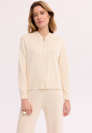 Kobieta ubrana w kremową bluzę na zamek i pasujące spodnie, stoi na jednolitym jasnym tle, patrząc prosto przed siebie.