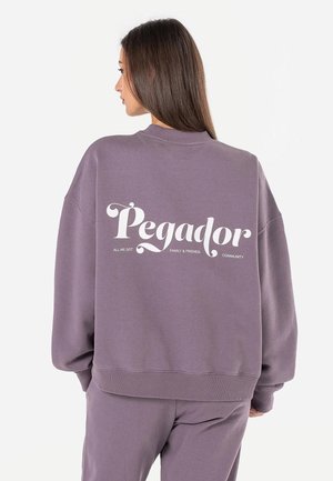 Kobieta o długich brązowych włosach, ubrana w luźną fioletową bluzę i spodnie, na plecach bluzy napis "Pegador" z mniejszymi napisami poniżej.