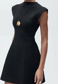 Robe noire sans manches avec un corsage ajusté et une jupe évasée ; fabriquée à partir d'un tissu texturé ; ornée d'un collier pendentif en feuille d'or.