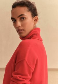 Red turtleneck sweater with ribbed collar and cuffs, featuring a soft texture. Model has short hair and small hoop earrings.