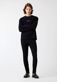 Sweatshirt noir avec logo discret, associée à un pantalon noir ajusté et des chaussures noires épaisses. Le modèle pose les bras croisés devant un fond uni.