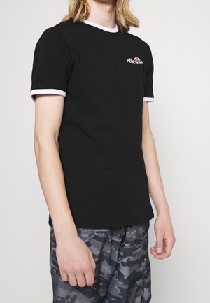 Osoba ubrana w czarną koszulkę z białym wykończeniem i logo Ellesse, w połączeniu z szaro-czarnymi spodniami w kamuflażowy wzór, stojąca na jednolitym tle.