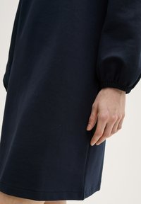 Abito blu navy realizzato in tessuto testurizzato. Presenta una vestibilità ampia, maniche lunghe con polsini elasticizzati e un orlo dritto. Una mano è parzialmente visibile.