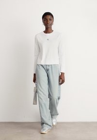 T-shirt bianco a maniche lunghe con un piccolo logo blu, abbinato a jeans chiari e morbidi, e sneaker argentate. Borsa semplice argentata.