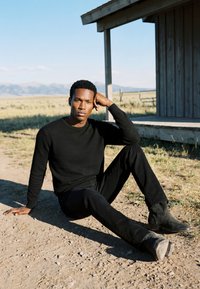 Homme portant un pull noir et un pantalon assis sur le sol en terre près d'une véranda en bois dans un paysage rural ouvert sous un ciel clair.