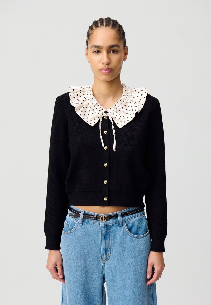 Cárdigan negro recortado con un cuello de volantes de lunares crema y cierre frontal de botones, combinado con jeans de tiro alto en azul claro.