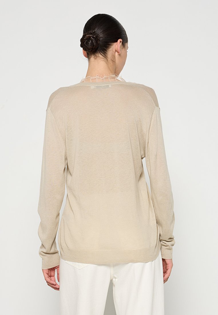 Beige langemouwen trui met een relaxte pasvorm, lichtgewicht breimateriaal en subtiele rafelige details bij de halslijn, casual gedragen.