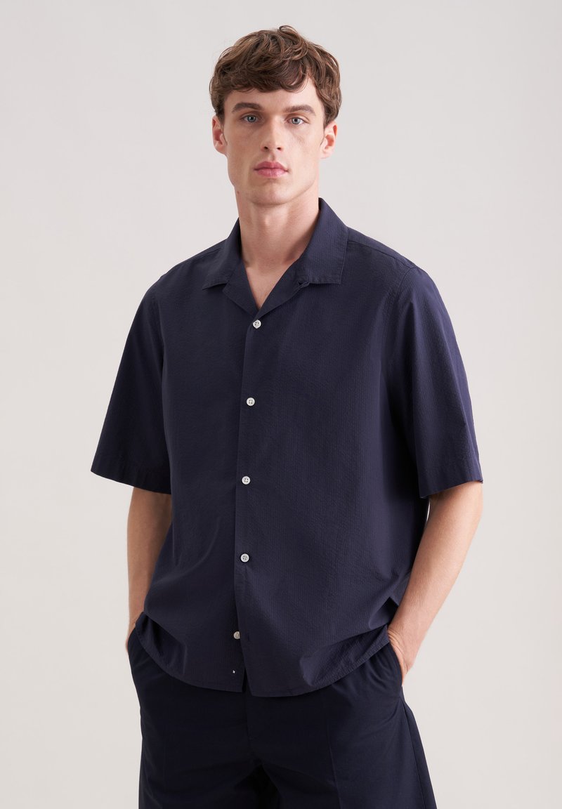 Jeune homme portant une chemise bleu marine à manches courtes et un pantalon bleu marine assorti, debout les mains dans les poches devant un fond uni.