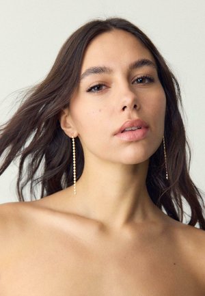 Mlada ženska z dolgimi temnimi lasmi, ki nosi dolge zlato verižne uhane in gleda rahlo navzgor na enobarvnem svetlem ozadju.