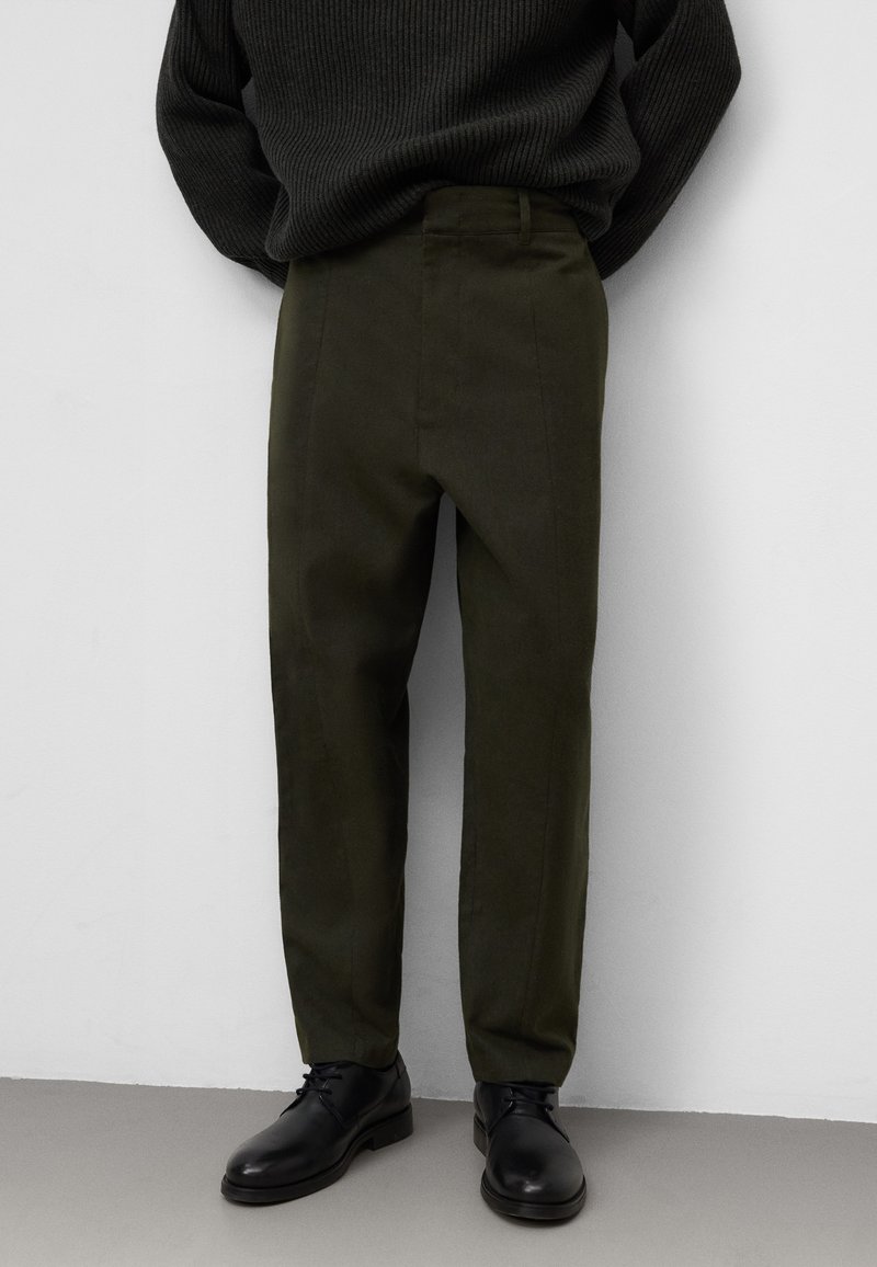 Olijfgroene op maat gemaakte broeken van een gestructureerde stof, met een vlakke voorkant, zijzakken en een slim fit. Gecombineerd met zwarte schoenen.