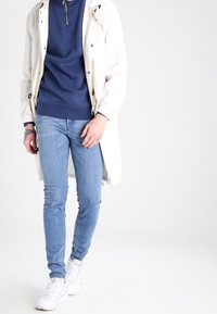 White long jacket over a navy sweatshirt, with light blue skinny jeans and white sneakers. The outfit features casual, simple design.