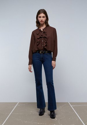 Brown, long-sleeve blouse with ruffles down the center, button closure, paired with dark flared jeans and black ankle boots.