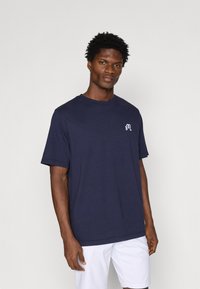 Jūras zilas kokvilnas t-krekls ar īsām piedurknēm, apaļu kakla izgriezumu un mazu baltu logotipu uz krūtīm. Gluda tekstūra, brīvs piegriezums un ikdienišķs stils.