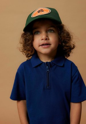 Granatowa koszulka polo z zamkiem błyskawicznym, w połączeniu z zieloną czapką z pomarańczowym logo drużyny futbolowej i numerem 85.