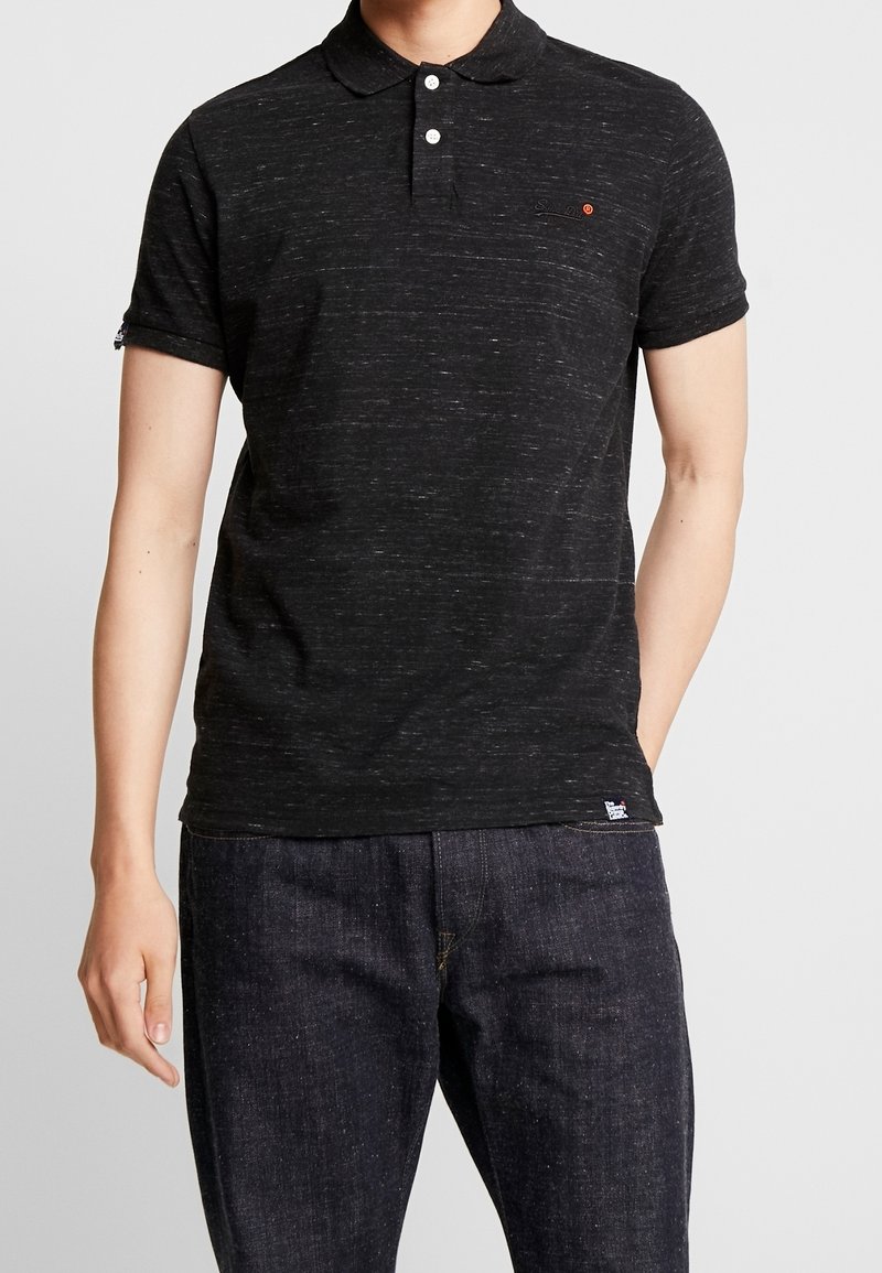 Polo shirt noir à motifs tachetés avec un plastron à trois boutons, des manches courtes, un petit logo sur la poitrine, et un tissu texturé. Associé à un jean foncé.