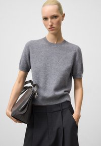 Maglione grigio a maniche corte in maglia con collo rotondo, abbinato a pantaloni scuri formali e una borsa in pelle scura. Texture liscia, design minimale.