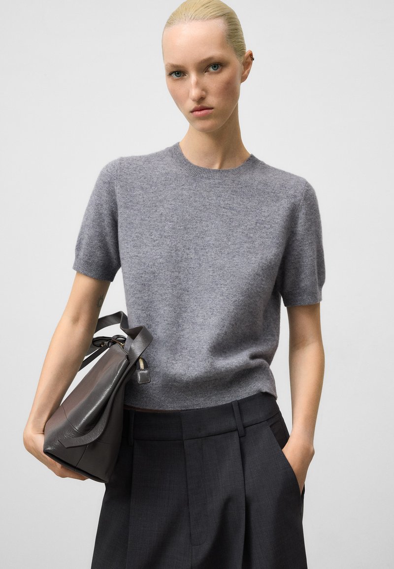 Maglione grigio a maniche corte in maglia con collo rotondo, abbinato a pantaloni scuri formali e una borsa in pelle scura. Texture liscia, design minimale.