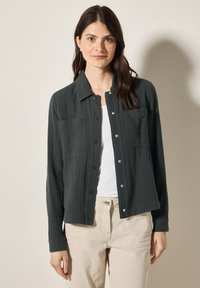 Camicetta grigia scura con texture, con bottoni nella parte anteriore, due tasche sul petto e un colletto, indossata sopra un top bianco e pantaloni beige.