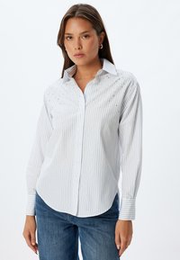 Camicia bianca con righe verticali azzurre e decorazioni sulle spalle. Presenta un colletto classico e maniche lunghe.