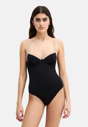 Donna con lunghi capelli scuri che indossa un costume intero nero con spalline color nude, in piedi davanti a uno sfondo chiaro e uniforme.