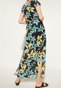 Maxikleid aus schwarzem, durchscheinendem Stoff mit floralem Muster in Türkis- und Gelbtönen, kurzen Ärmeln und einem seitlichen Schlitz.