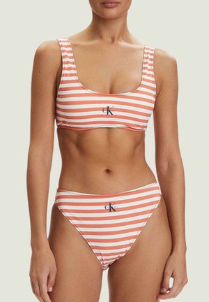 Mujer con bikini a rayas naranja y blanca con logotipos de Calvin Klein en la parte superior e inferior sobre un fondo neutro.