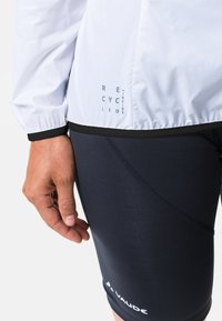 Weiße, leichte, recycelte Jacke mit elastischem schwarzem Saum, gekennzeichnet mit "RECYCLED" in Blau, kombiniert mit enganliegenden schwarzen Radfahrshorts.