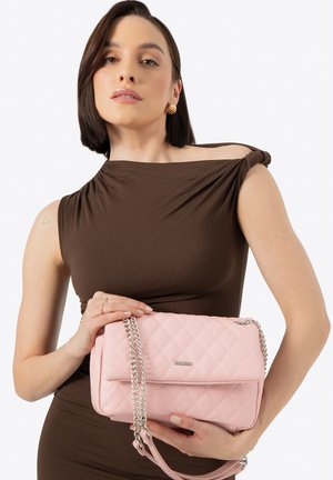 Rosa gesteppte Handtasche mit Kettenriemen, mit glatter Textur und einem Metalllogo. Wird von einer Person gehalten, die ein braunes Kleid trägt.