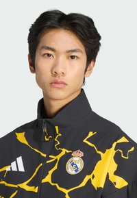 Musta takki, jossa keltainen salamanisku-kuvio, vetoketjullinen kaulus ja Real Madridin logo rinnassa; valmistettu hengittävästä materiaalista.