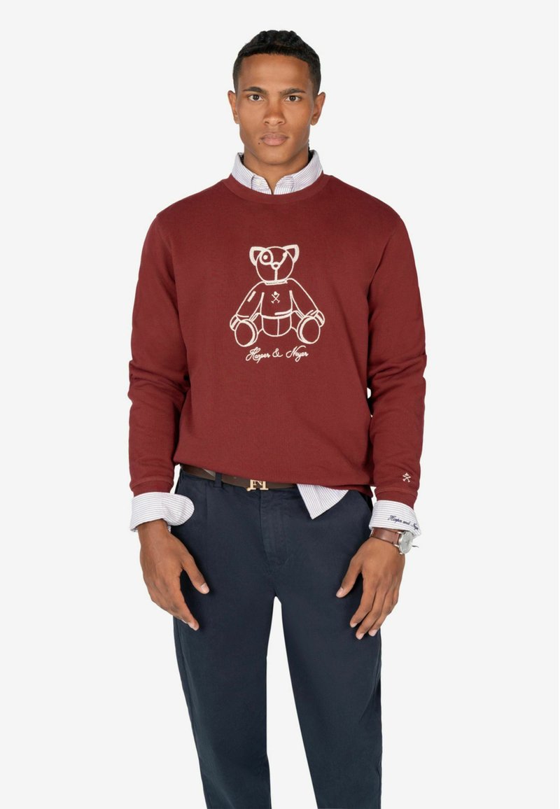 Bordeauxfarbener Pullover aus Baumwolle mit einem Grafikdesign und Logo eines weißen Bären. Kombiniert mit navyblauen Hosen und einem geschichteten Hemd.