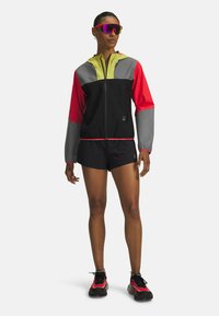 Farbenfrohes Sportoutfit mit einer schwarzen Jacke, die rote, graue und neongelbe Akzente hat, kombiniert mit schwarzen Shorts und sportlichen Schuhen.