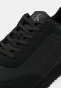 Baskets noires Calvin Klein avec lacets noirs, couture texturée et logo « cK » visible sur la languette et le panneau latéral.