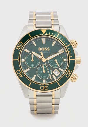 Men’s silver and gold metal wristwatch with green bezel, green dial, three subdials, date window, and gold-tone hour markers and hands.