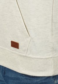 Jasno beżowy hoodie z kangurzą kieszenią i teksturowanym materiałem. Zawiera małą brązową skórzaną metkę z detalem marki na bocznej szwie.