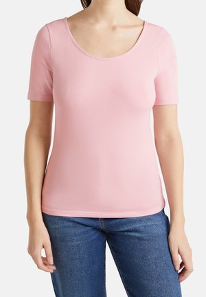 Femme portant un t-shirt rose clair ajusté à manches courtes et col rond, et un jean en denim bleu, debout devant un fond uni.